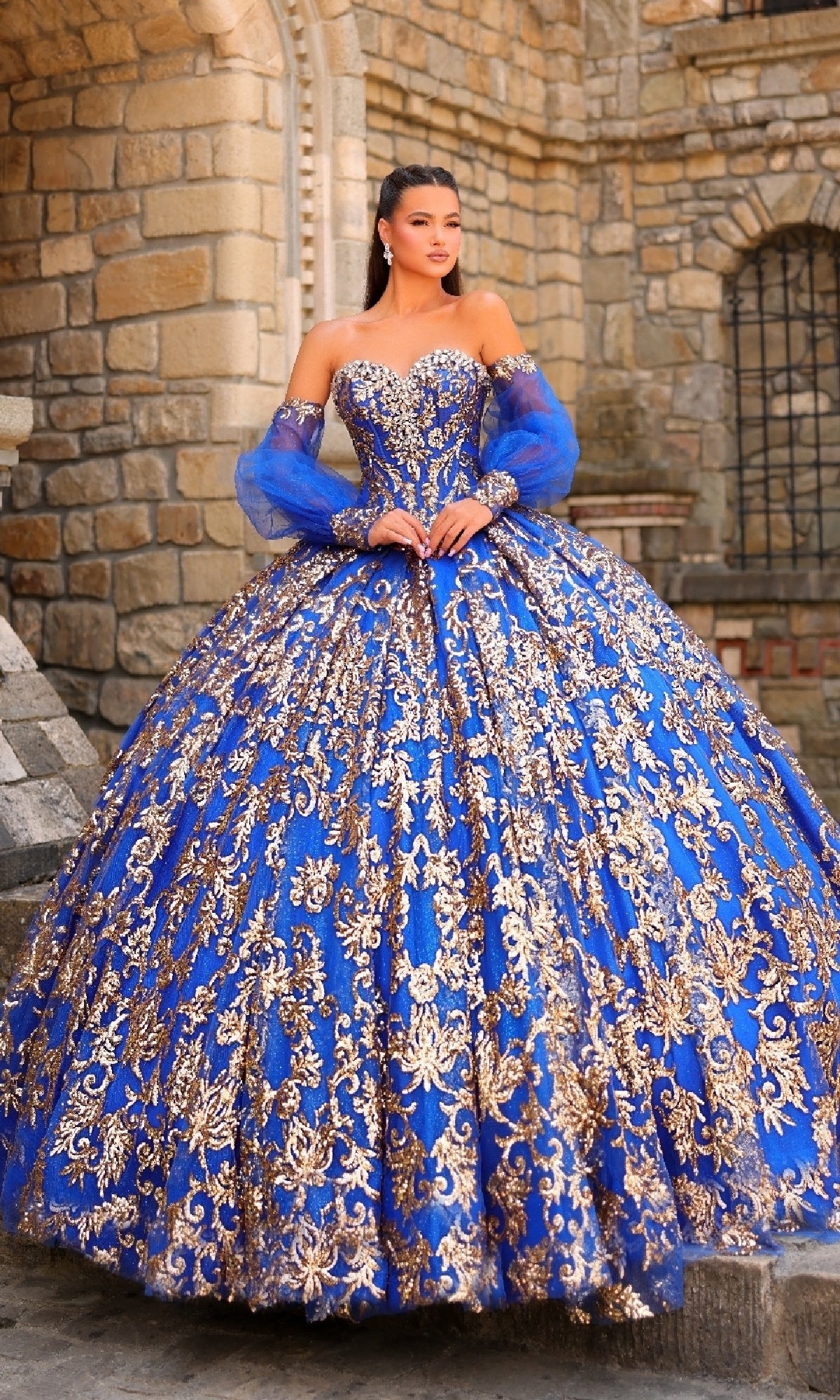 A woman stands in front of a stone building wearing the Sequin Quinceanera Dress: Amarra 54238, a blue ball gown featuring gold embellishments, a corset bodice, and sheer blue sleeves.